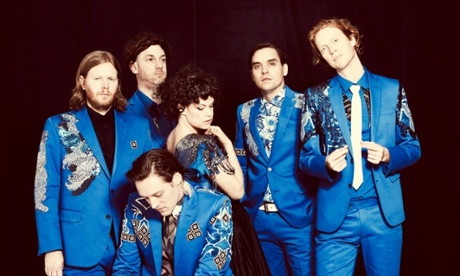 Arcade Fire band group shot against a black background, all wearing bright blue suits with applique patchwork.