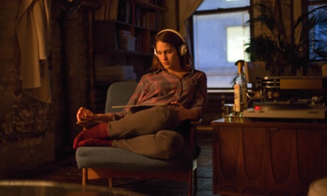 Lola Kirke as Tracy sits on an upholstered chair with her legs tucked up and to the side, in the warm glow of a lamp as she listens on headphones to a record playing on the turntable.