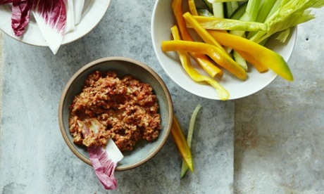 A pottery dish of muhamara, a blended dip of walnuts and red peppers, on a marble tabletop with bowls of freshly cut raw salad vegetables.