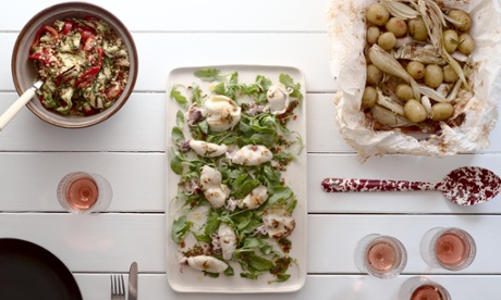 A rectangular dish of barbecued squid with a salad of fregola, grilled vegetables and basil, potatoes and fennel in a bag. On a white wooden table with wine glasses and a marbled serving spoon.