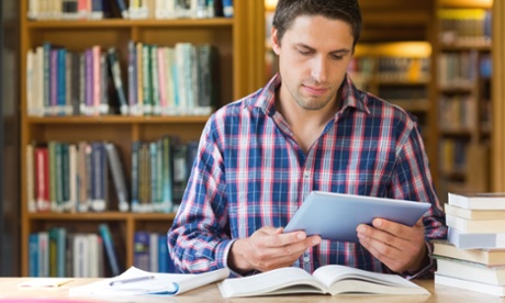 Man holding an iPad sitting at a desk surrounded by books in a library