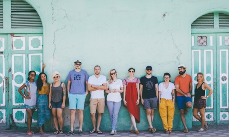 Group of travellers stand, moodily, against a wall. 