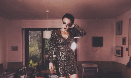 Actor Jess Latowicki stands on a kitchen table with heavy, black eyeliner, wearing a short sparkling dress