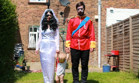 A man and a woman walk a toddler towards camera in a suburban garden. The man is dressed in a fake, red aristocratic dress uniform, the woman in a wedding dress