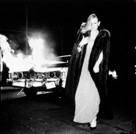 Patti Hansen, in finery of gown, fur coat and hair do, walks away from a burning car. This is glamour augmented with cinematic drama.