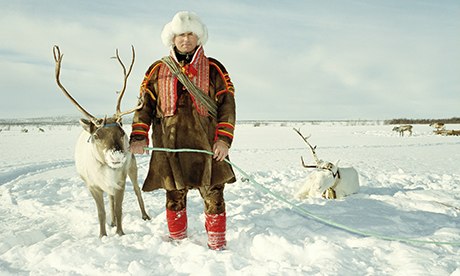 Reindeer herdsman with fur hat holds a reindeer by a long rope