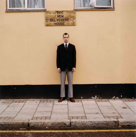 Isolation: A man stands very formally in an empty street, his back to the wall