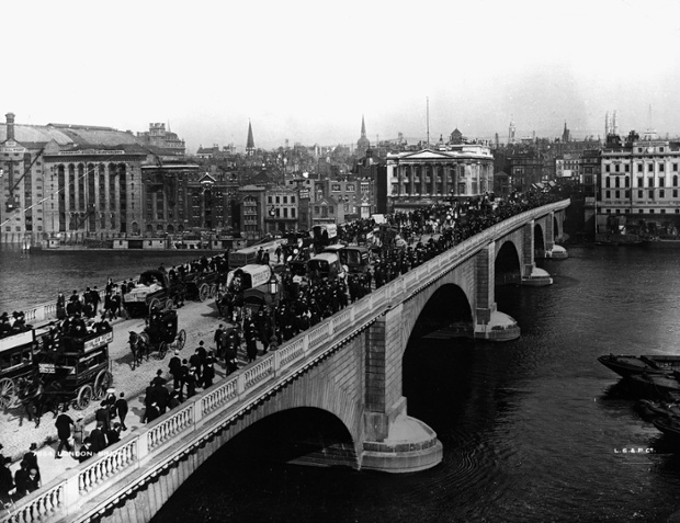Traffic on London Bridge over the river Thames in 1904. The bridge is full of pedestrians, horse drawn omnibuses and horse drawn carriages. Various wharfs and warehouses are visible on the left of the shot across the river.