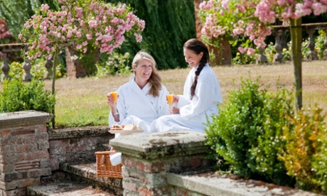 Two smiling women wearing white dressing gowns, holding glasses of orange juice in a pretty garden 